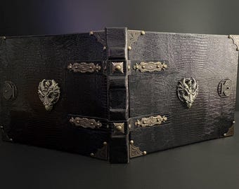 May include: A black, faux-leather book with a textured, reptilian pattern. The book features ornate metal corner protectors, decorative clasps, and dragon head embellishments. The spine is also black and has a raised design.