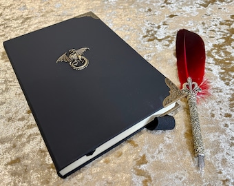 May include: A black leather-bound journal with a dragon design on the front cover. A red feather quill pen with a silver ornate handle is lying next to the journal.