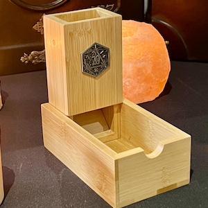 May include: A wooden dice tower with a metal D20 die on the front. The tower has a drawer on the bottom and a slot for dice to fall out of the top.
