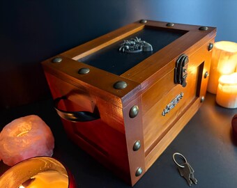 May include: Wooden chest with brass accents, a black top, and a decorative metal emblem. The chest has a handle and is secured with a latch. Decorative brass studs adorn the corners. A key and candles are in the background.
