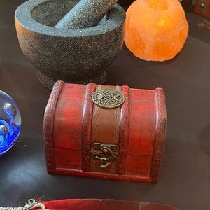 Op de afbeelding: Een kleine houten schatkist met een bruine leren band en een metalen sluiting. De kist heeft een decoratief metalen ontwerp op de deksel. De kist staat op een donker oppervlak.