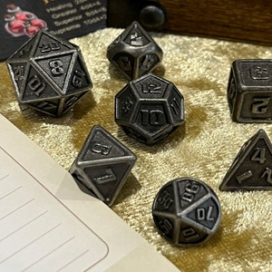 May include: A set of seven metal dice in various shapes and sizes. The dice are silver in color and have black numbers. The dice are on a gold-colored surface.