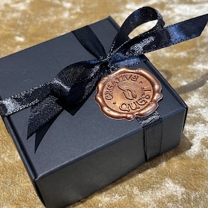 Può includere: Una scatola regalo nera con un nastro e un fiocco neri. Un sigillo di cera color rame con il testo "Creative Quest" è sulla parte superiore della scatola.