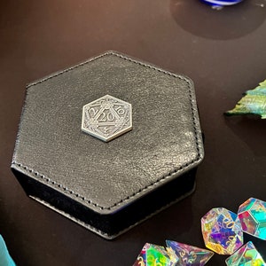Può includere: Una scatola da gioco esagonale nera con un emblema D20 in metallo argentato sul coperchio. La scatola è circondata da dadi iridescenti.