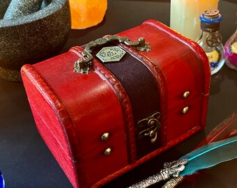 May include: A small, red wooden treasure chest with a metal latch and handle. The chest has a brown leather strap with a decorative metal emblem on the top.