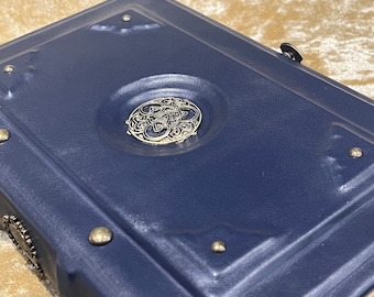May include: A dark blue leather-bound journal with a silver Celtic knot design on the front cover. The journal has metal accents and a decorative clasp.