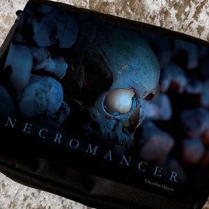 May include: A black messenger bag featuring a blue and white skull graphic with the text "NECROMANCER CREATIVE QUEST".