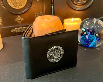 May include: A black leather wallet with a silver Celtic knot design on the front. The wallet is closed and sitting on a dark surface.