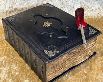 May include: A black leather-bound journal with gold accents and a bee design on the cover. A silver pen with a red feather quill is resting on the journal.