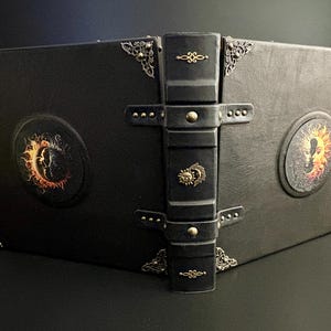 Op de afbeelding: Een zwart leren boek met decoratieve metalen hoekaccenten en een centrale sluiting. De omslag heeft cirkelvormige ontwerpen met een zon- en maanmotief in oranje en geel. De rug heeft een decoratief metalen embleem.