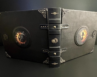May include: A black leather-bound book with decorative metal corner accents and a central clasp. The cover features circular designs with a sun and moon motif in orange and yellow. The spine has a decorative metal emblem.
