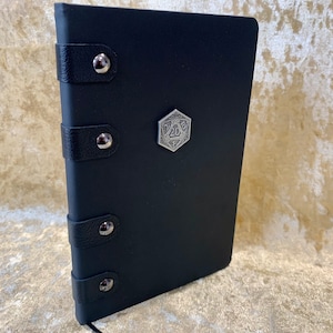 May include: Black leather-bound journal with a silver metal D20 die on the front cover. The journal has four silver metal rivets on the spine.