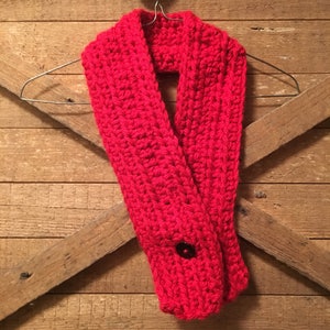 May include: A vibrant red, hand-crocheted scarf with a button closure. The scarf is displayed on a wire hanger against a rustic wooden background. The chunky knit design and bold color make it a cozy winter accessory.