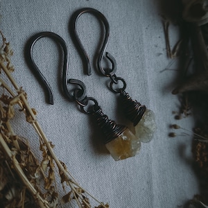 May include: A pair of earrings with a raw, yellow-brown crystal wrapped in copper wire. The earrings are attached to a simple, dark metal hook.