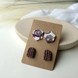 May include: A set of three pairs of polymer clay earrings. Two pairs are hexagonal shaped with abstract patterns in shades of pink, white, gray, and orange. The third pair is a brown, textured, arched shape.