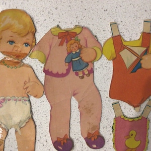 May include: A vintage paper doll set featuring a blonde doll with a pink dress, a yellow and orange dress, and a pink bib. The doll is holding a smaller doll in a blue dress.