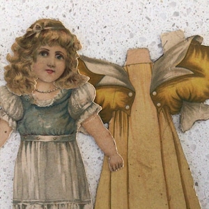 May include: A vintage paper doll with blonde hair wearing a blue and white dress, positioned next to a separate yellow dress with large puffed sleeves. The doll features a detailed face and a necklace, while the yellow dress has visible paper tabs.