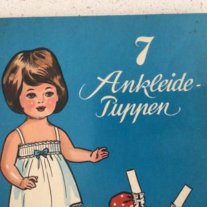 May include: A vintage illustration of a doll with brown hair and blue eyes, wearing a white dress with a blue bow. The number 7 and the words "Ankleide-Puppen" are written in white script on a blue background. The image is a vintage paper doll.
