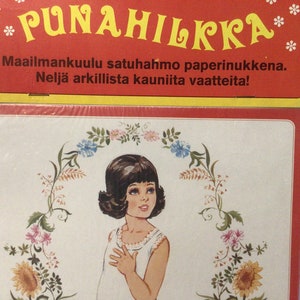 Puede incluir: Un conjunto de muñecas de papel vintage con una niña de pelo castaño y un vestido blanco. La muñeca está rodeada de una corona de flores con girasoles, flores rosas y azules. El embalaje presenta el texto "Punahilkka" en letras amarillas sobre fondo rojo. El texto "Maailmankuulu satuhahmo paperinukkena. Neljä arkilista kauniita vaatteita!" también está incluido en el embalaje.
