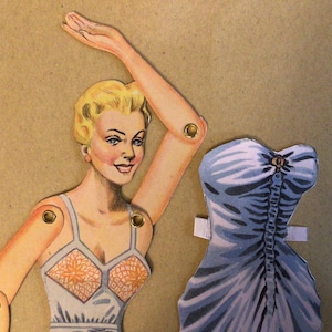 May include: A vintage paper doll with blonde hair, wearing a light-coloured top with orange floral patterns, featuring articulated limbs connected by metal brads. Beside the doll is a separate blue strapless dress with a central button detail.