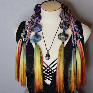 May include: A mannequin head displays colorful hair extensions in shades of purple, blue, green, yellow, and red. The extensions are styled with bulbous shapes and long, rainbow-colored strands. A black top and a pendant necklace complete the look.