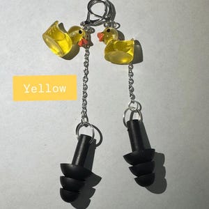 May include: A pair of earplugs attached to silver-toned metal chains and small yellow translucent rubber duck charms. The earplugs are black with a three-tiered flange design. A yellow rectangular graphic with the text Yellow is visible.