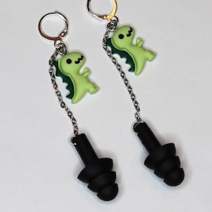 May include: A pair of black silicone earplugs with a silver chain attached to a green dinosaur charm. The charms are attached to silver hoop earrings.