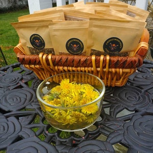 May include: A wicker basket filled with brown paper bags labeled "A Whole Mood Shop's Herbal Boutique". The basket is sitting on a black metal table with a floral design. A glass bowl filled with yellow dandelion flowers is in front of the basket.