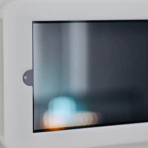May include: A white wall-mounted tablet frame with a black screen. The screen is reflecting light from a window.