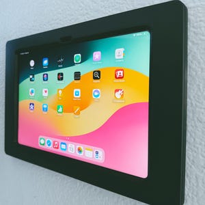 May include: A black wall mount frame with a tablet inside. The tablet screen displays a variety of colorful app icons on a pink and yellow background.