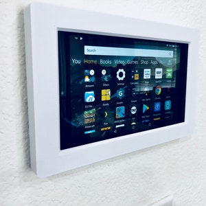 May include: A white framed tablet mounted on a white wall. The tablet screen displays a variety of apps, including Amazon, Google, and Minecraft. The search bar at the top of the screen reads "Search".