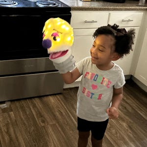 Op de afbeelding: Een gele poppenkop met een oplichtende functie, vastgehouden door een persoon. De pop heeft een paarse neus, een roze mond en een grijze handschoen. De persoon draagt een grijs t-shirt met de tekst "FAVO ITE SISTER".