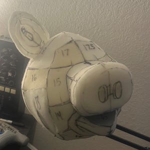 Peut inclure: Une tête de cochon en mousse de couleur claire avec des marques et des chiffres au crayon. La tête de cochon a de grandes oreilles, un museau arrondi et est divisée en sections avec des lignes. Les chiffres sont écrits au crayon sur la mousse.