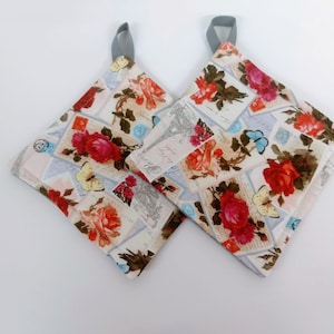 Könnte beinhalten: Ein Paar weiße Topflappen mit einem Blumenmuster aus roten Rosen, Schmetterlingen und Vintage-Postkarten. Die Topflappen haben graue Schlaufen zum Aufhängen.