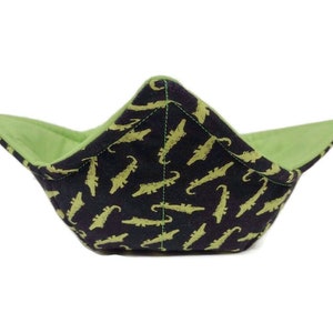 May include: A fabric bowl cosy with a black exterior patterned with light green alligators. The interior is a solid light green. The cosy is designed to hold a bowl of hot food and protect the user's hands.