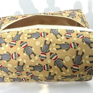 May include: A beige zippered pouch with a repeating pattern of sock monkeys and white buttons. The sock monkeys are gray with red and white accents. The pouch has a beige interior and a white zipper.