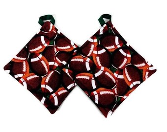 May include: A pair of brown and green pot holders with a pattern of footballs. The pot holders have a green loop handle.