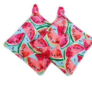 May include: Two square pot holders with a watermelon print. The pot holders are light blue with pink and black watermelons. The pot holders have pink fabric loops for hanging.