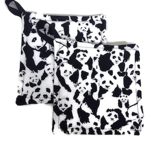 Puede incluir: Dos agarraderas cuadradas con un diseño de oso panda en blanco y negro. Una tiene un lazo negro para colgar. El patrón de panda se repite sobre el fondo blanco.