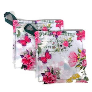 May include: Two white cotton pot holders with a floral print. The print features pink flowers, green leaves, and butterflies. The pot holders have a green ribbon attached to one of them. The text "Yonne Treilles Croissy" is visible on the pot holders.