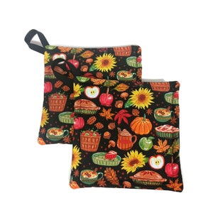 May include: Two black pot holders with a fall-themed print. The print features apples, pumpkins, pies, sunflowers, and leaves in shades of red, orange, yellow, and green.