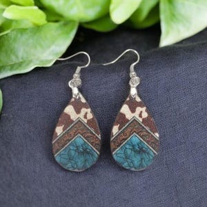 May include: A pair of teardrop earrings with silver hooks. The earrings feature a brown and white cowhide pattern at the top, a brown patterned center, and a turquoise blue bottom section. The earrings are displayed on a dark blue fabric.