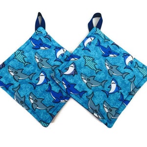 May include: Two blue and white pot holders with a shark pattern. The pot holders are made of fabric and have a loop for hanging.