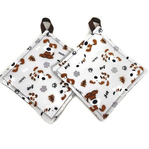Puede incluir: Dos agarraderas cuadradas con fondo blanco y un patrón repetido de perros de dibujos animados marrones y blancos, huellas de patas y las palabras "bark!" y "arf." Cada agarradera tiene un lazo marrón para colgar.