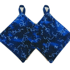 May include: A pair of blue pot holders with a constellation pattern. The pot holders are made of fabric and have a loop for hanging.