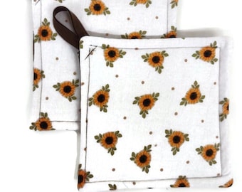 May include: Two white cotton pot holders with a sunflower and polka dot pattern. The pot holders have brown fabric loops for hanging.