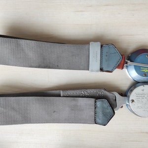 May include: Two gray seatbelts with metal buckles and circular mechanisms. The seatbelts have a woven texture and are laid out on a light-colored wooden surface. The mechanisms have blue and silver accents, with some text visible.
