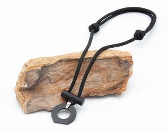 May include: A black paracord necklace with a black metal fire starter pendant. The pendant has a ring and a small bar. The necklace is displayed on a piece of petrified wood.