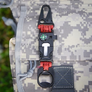 May include: A black multi-tool keychain with a compass, bottle opener, and a carabiner clip. The keychain is made of durable plastic and metal and has a red paracord strap.