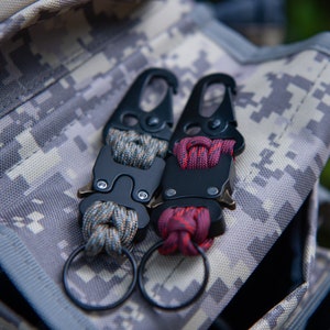 May include: Two black metal keychains with a loop and a clip. One keychain has a grey and brown paracord, and the other has a red and black paracord.